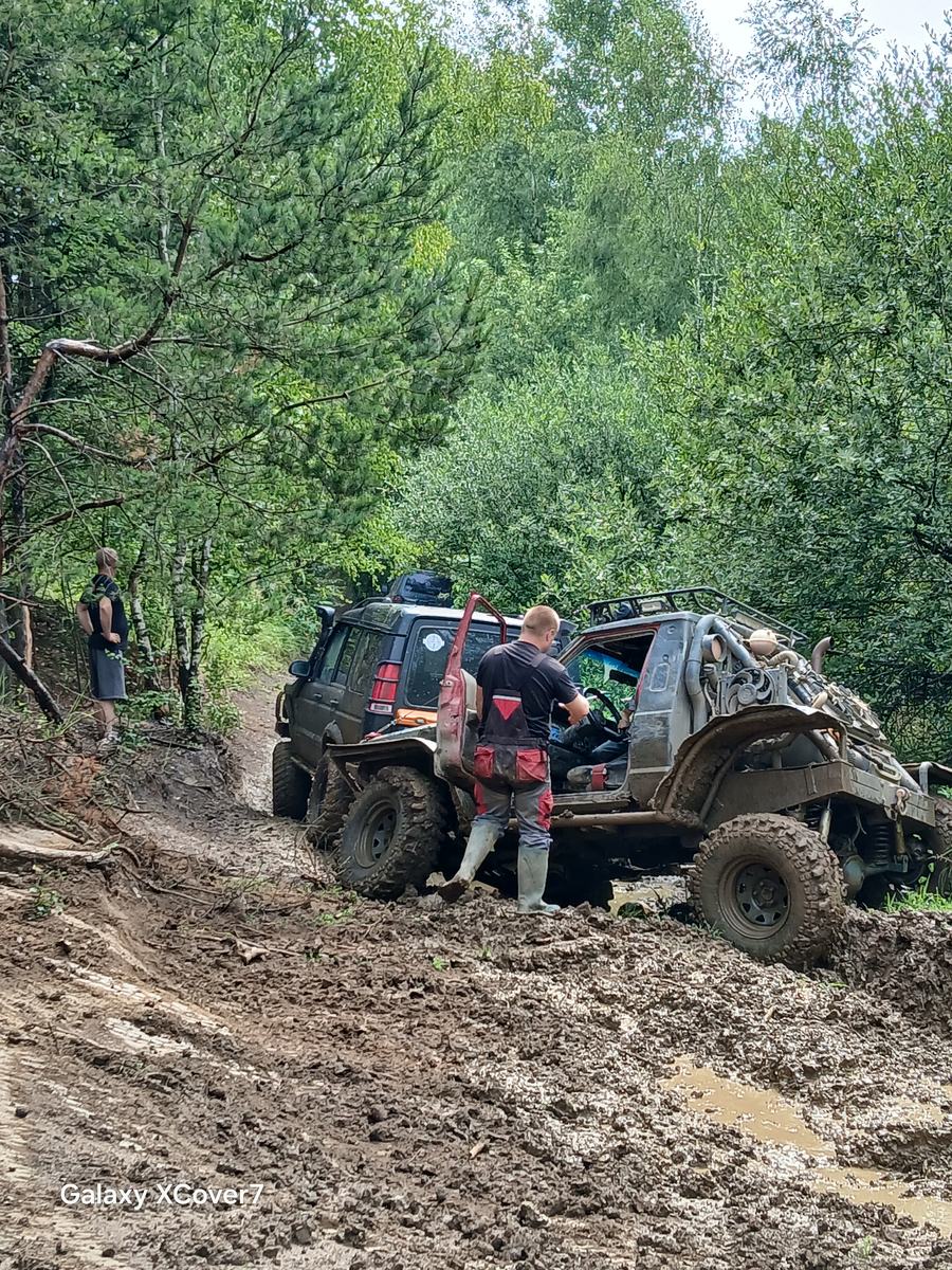 Dwa auta terenowe zakopane w błocie na leśnym odcinku toru, obok stoją uczestnicy